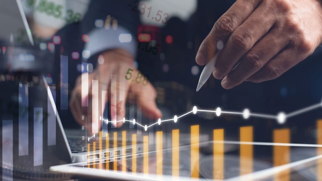 person standing over their laptop, holding a pen in the background, with graphs and line charts in low opacity in the foreground